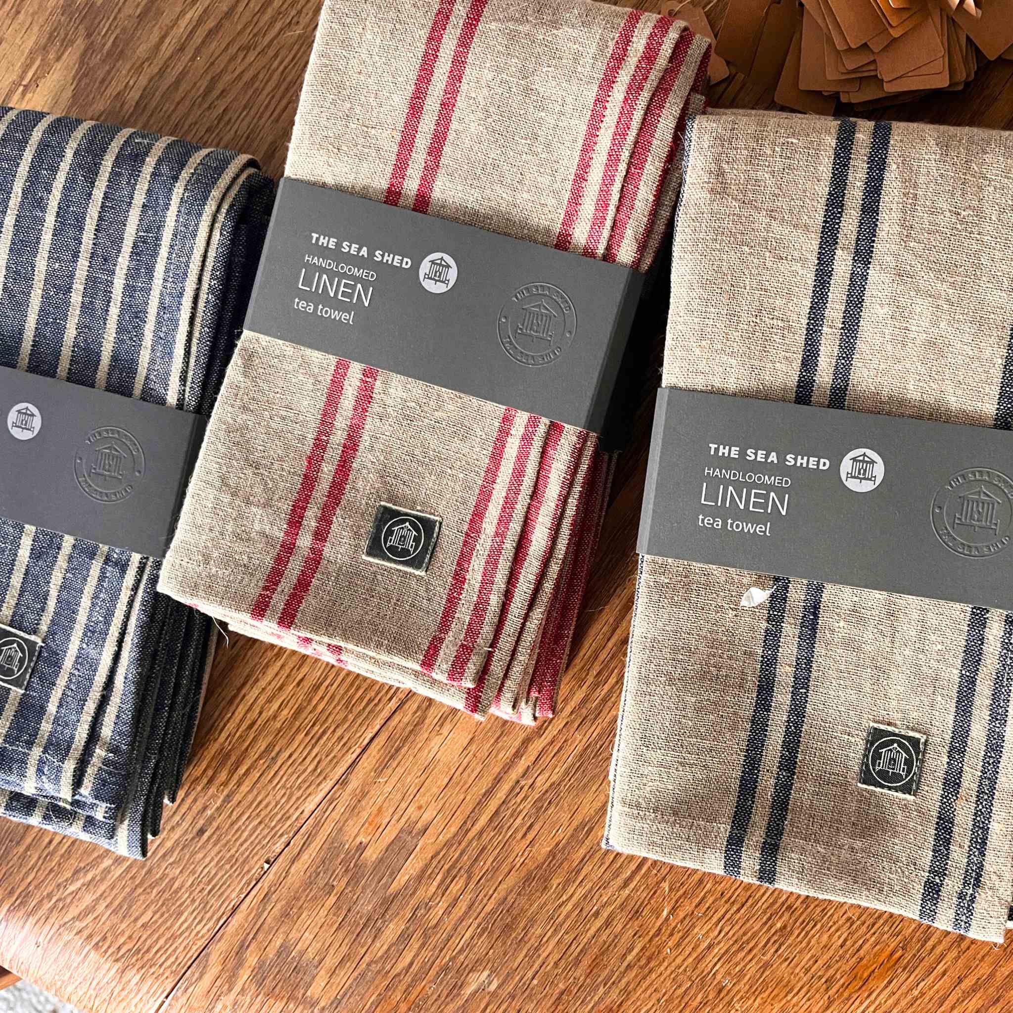 Three folded linen tea towels from The Sea Shed are arranged on a wooden surface. One towel has a blue and white stripe pattern, while the other two are beige with red and navy stripes.