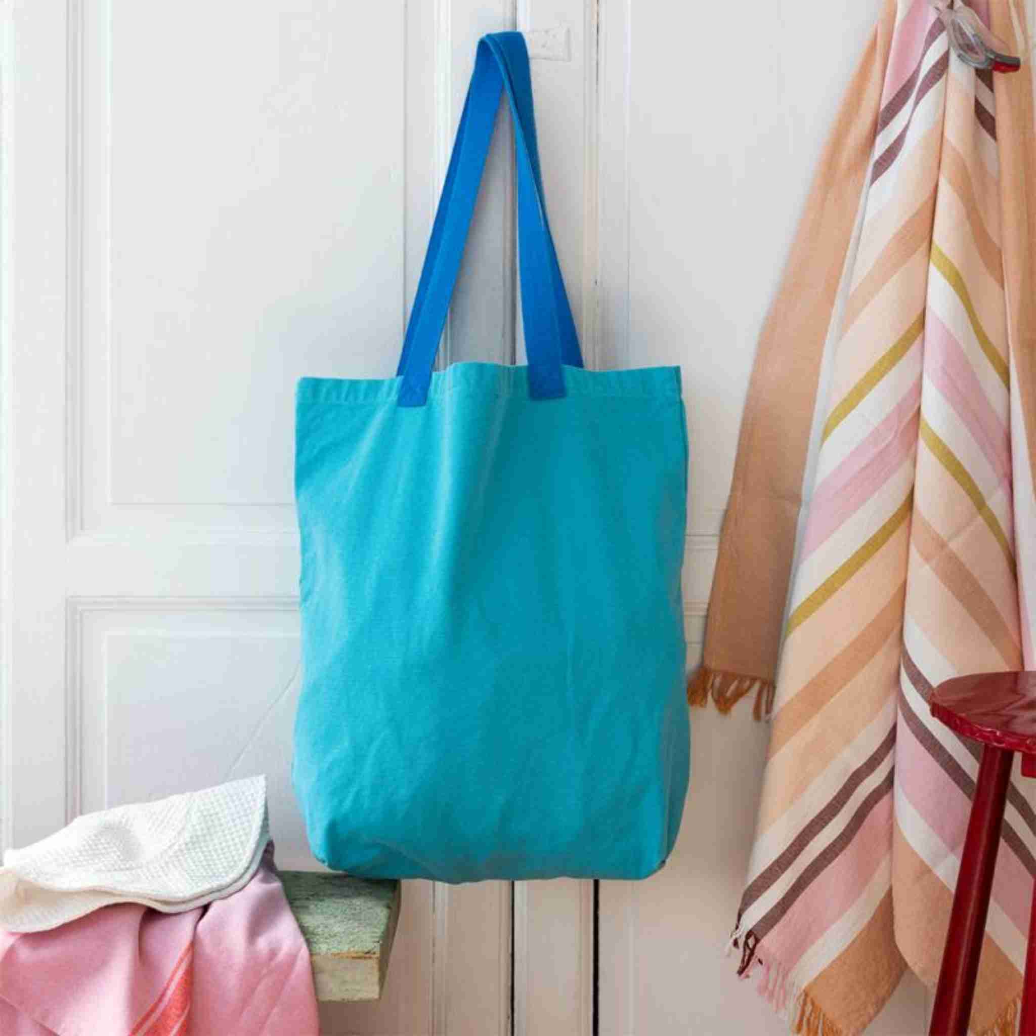 Turquoise tote bag hanging on a white wooden door with colorful towels in the background.