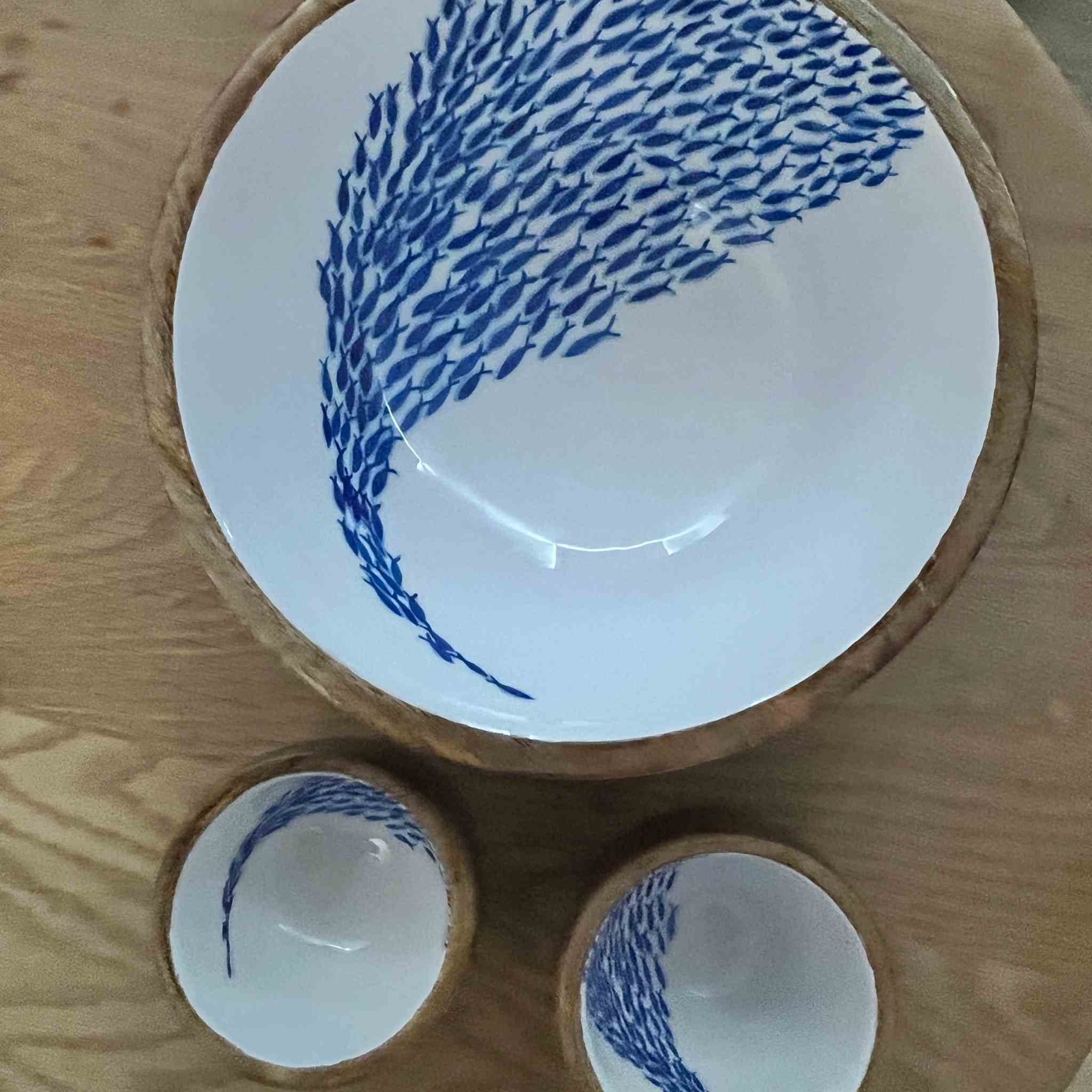A set of wooden tableware with blue fish-pattern designs and natural wood rims, including a large bowl and two smaller bowls, arranged on a wooden surface.