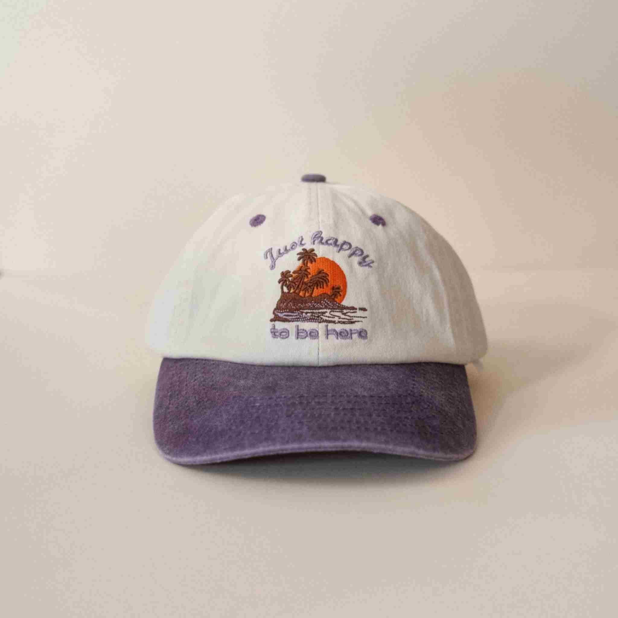 Cap with a beach scene and 'Happy to be here' text on a beige background