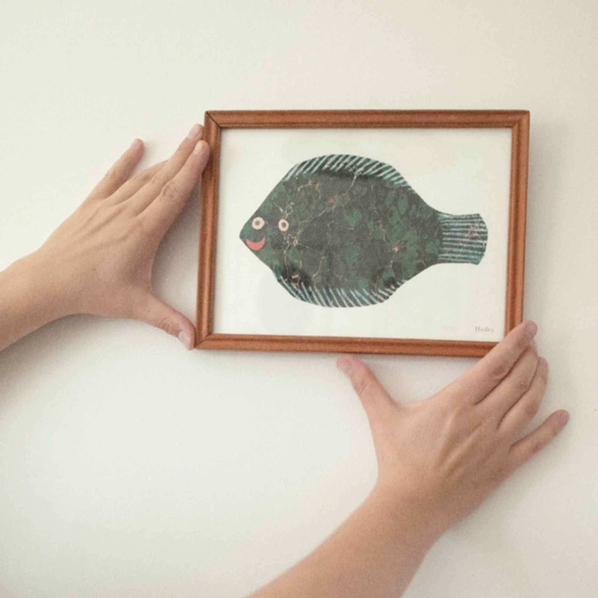 Framed artwork of a fish held by two hands on a white background