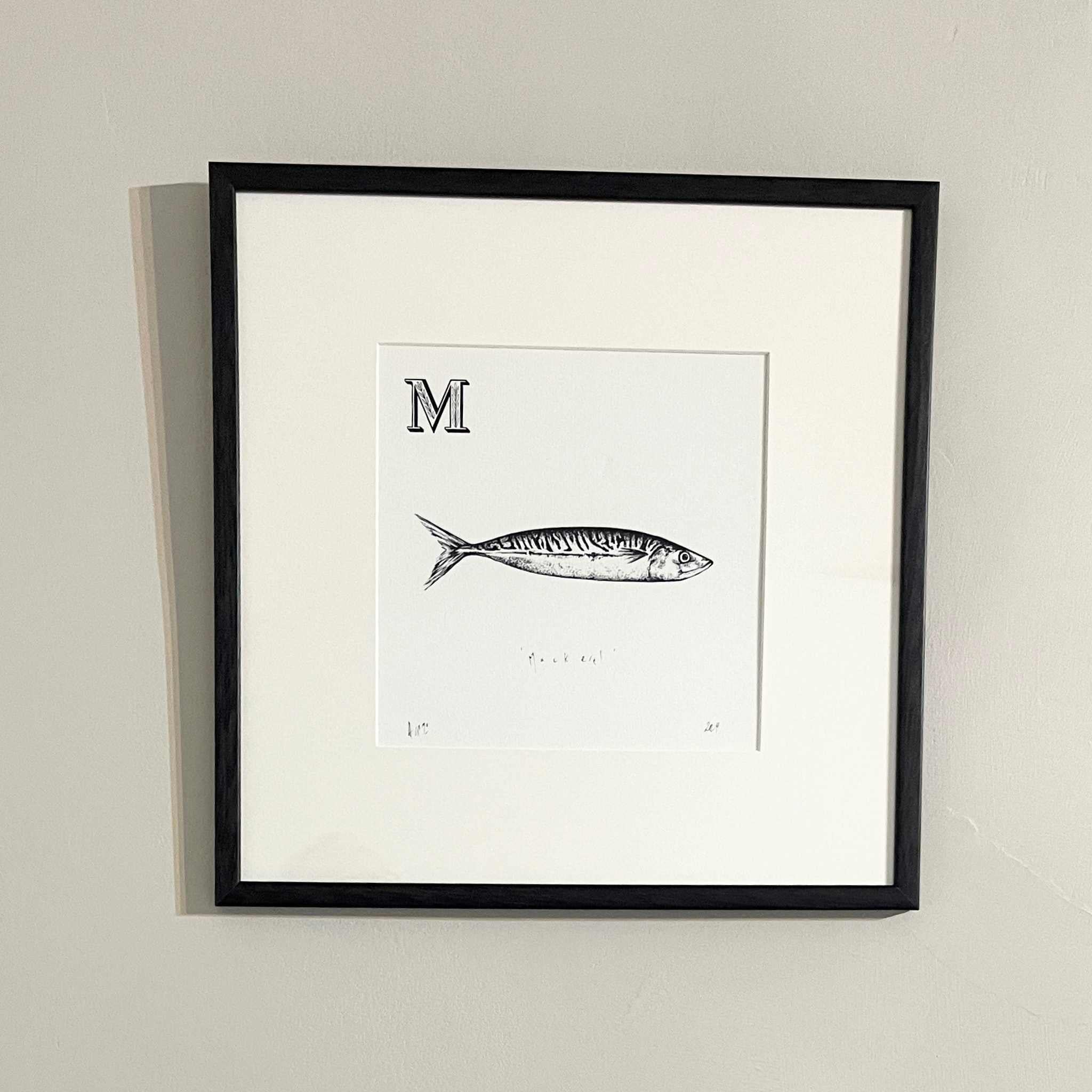 Framed black and white print of a fish illustration with letter 'M' on a beige wall.