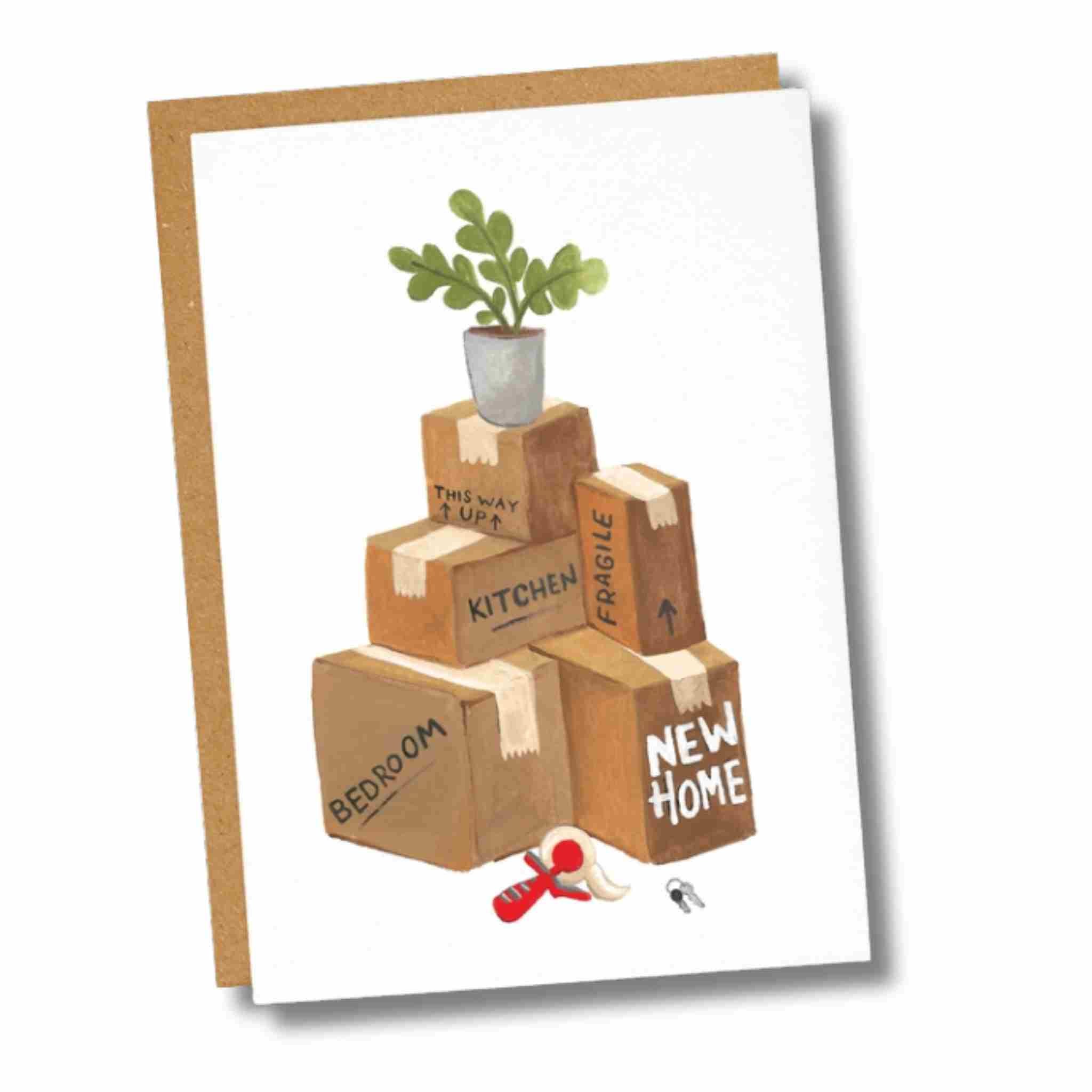 A square greeting card with a kraft envelope behind it, featuring an illustration of stacked cardboard boxes with a small potted plant on top. The boxes are labeled with words like BEDROOM, KITCHEN, FRAGILE, and NEW HOME, with a red tape measure and a small key at the bottom.