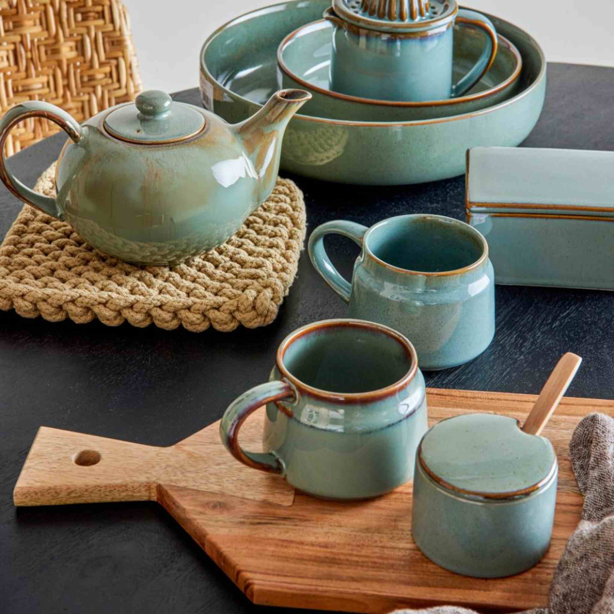 Set of ceramic teaware including teapot, mugs, and sugar bowl on a wooden board with a woven mat.