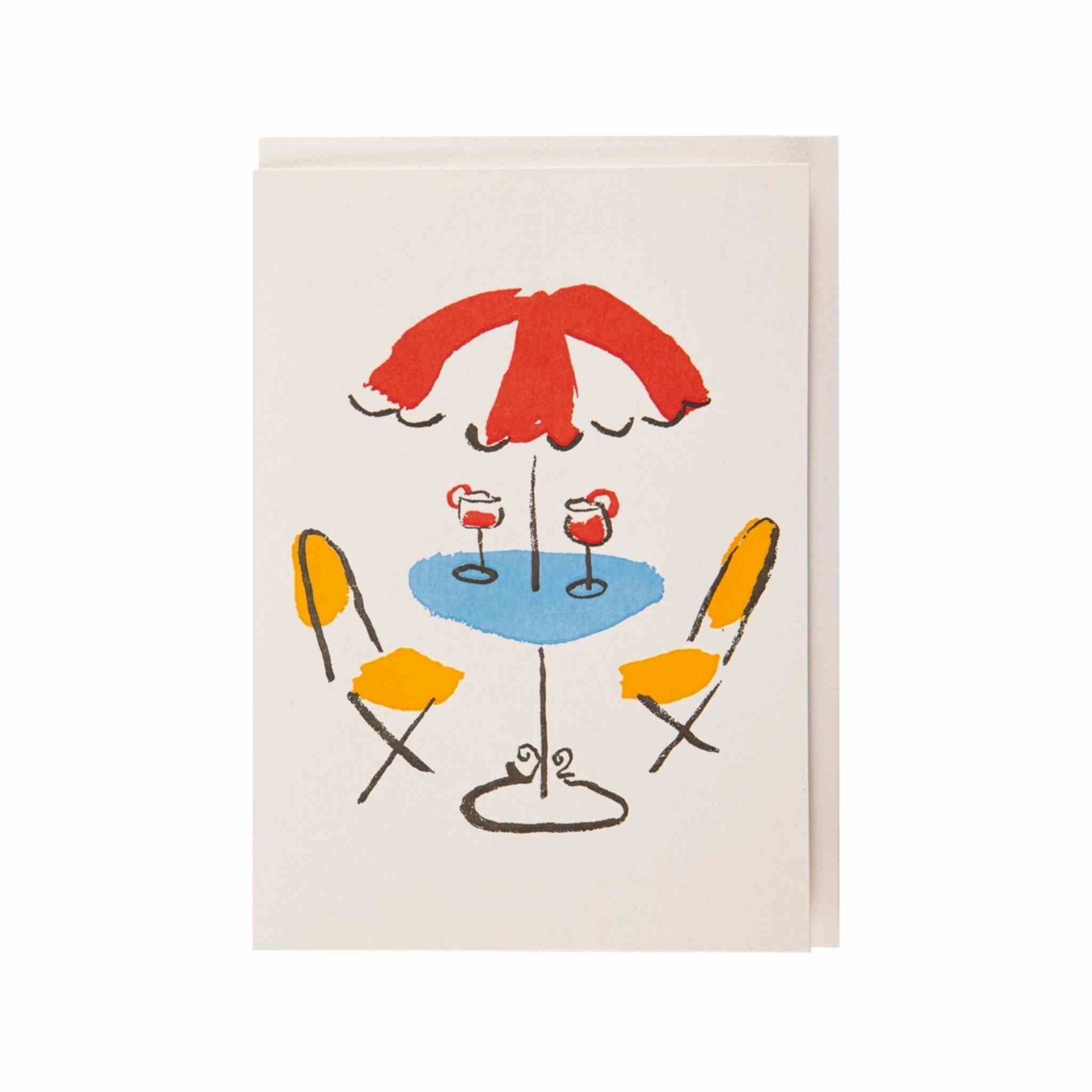 Agreeting card featuring a minimalist, hand-drawn style illustration of an outdoor cafe scene with a red umbrella, blue table, yellow chairs, and two cocktails, shown with a matching cream envelope behind it