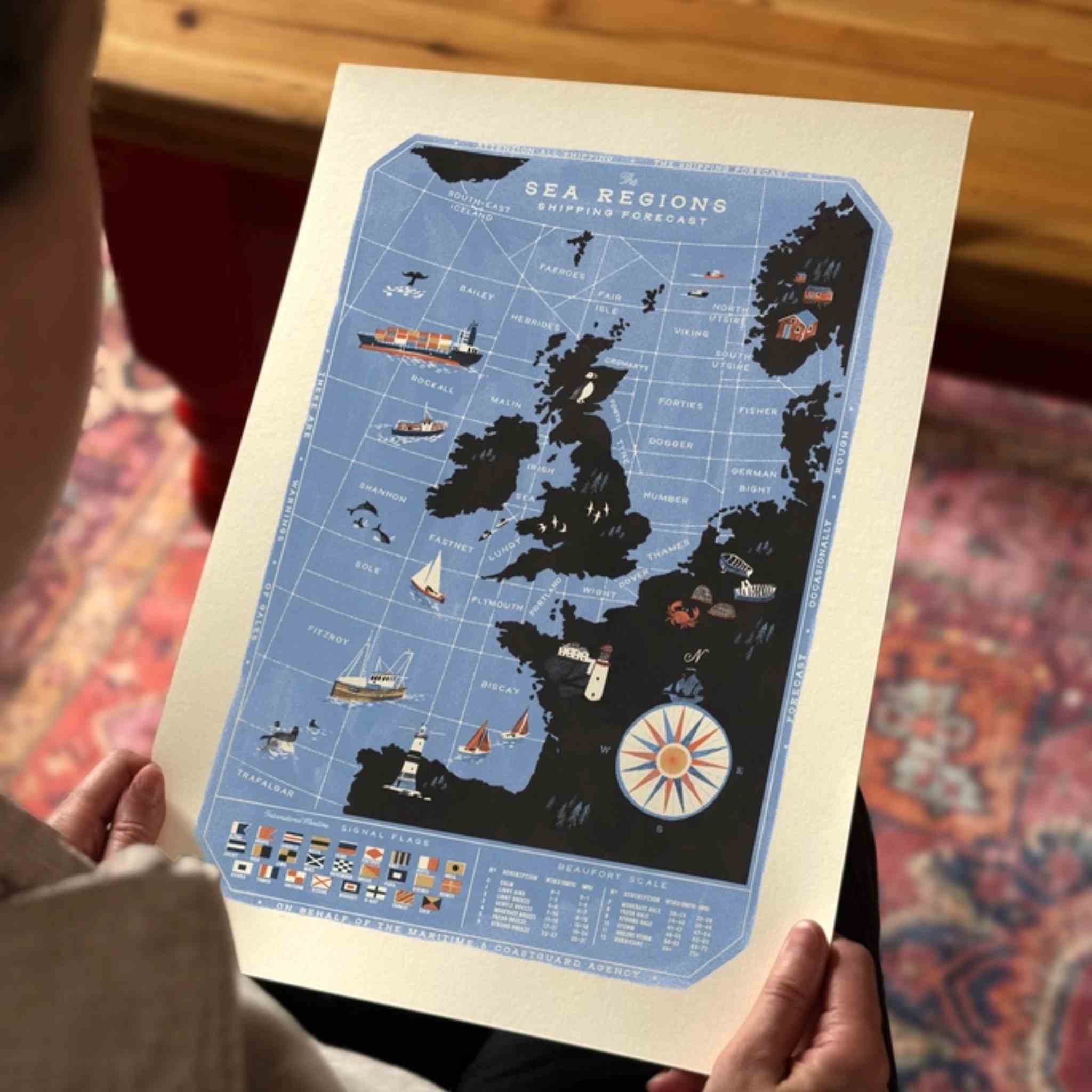 A person holds a vintage-style illustrated poster showing a blue sea regions map with black landmasses and nautical illustrations.