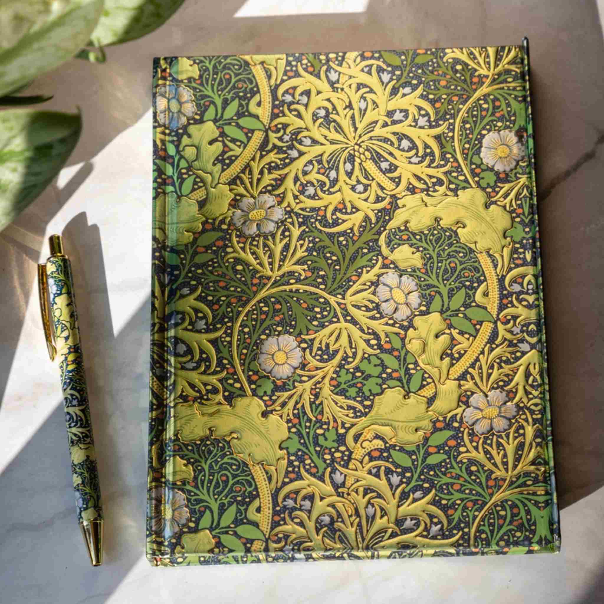 A hardcover notebook with a William Morris-style floral pattern in olive green, yellow, and cream, shown next to a matching patterned pen on a light marble surface with natural shadows.