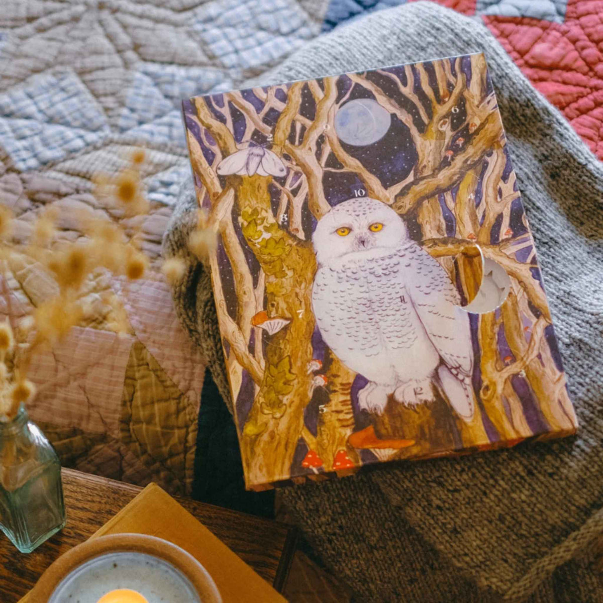 An art print featuring a white owl perched among gnarled tree branches under a full moon, styled on a cozy gray knit blanket with dried flowers and a candle nearby.