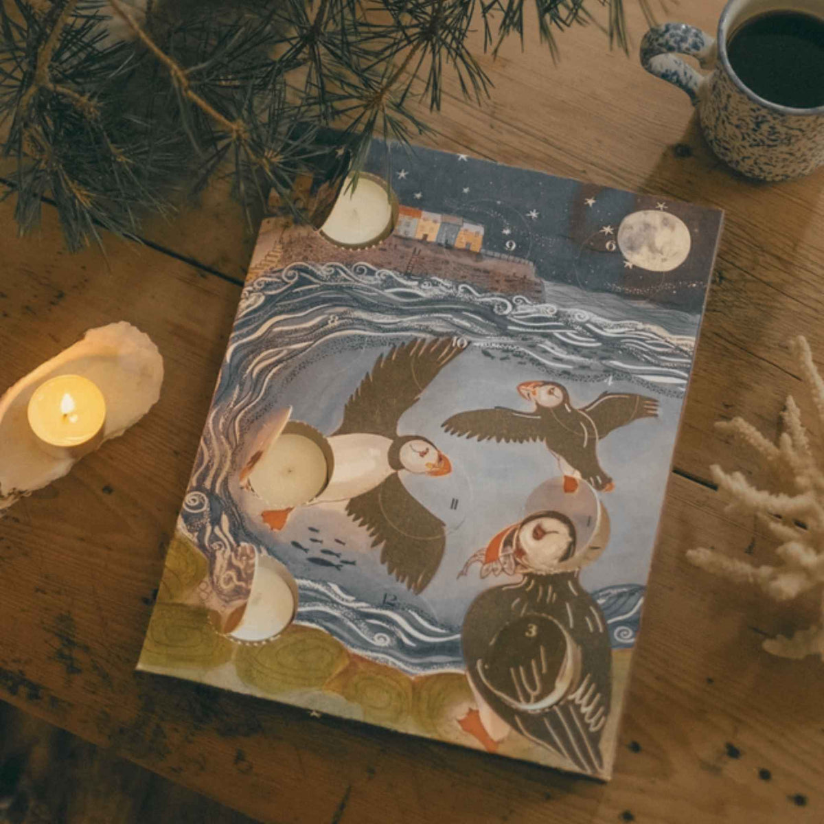An art print featuring a whimsical illustration of puffins in a moonlit lake, styled on a rustic wooden table with candles, pine branches, and a ceramic mug.