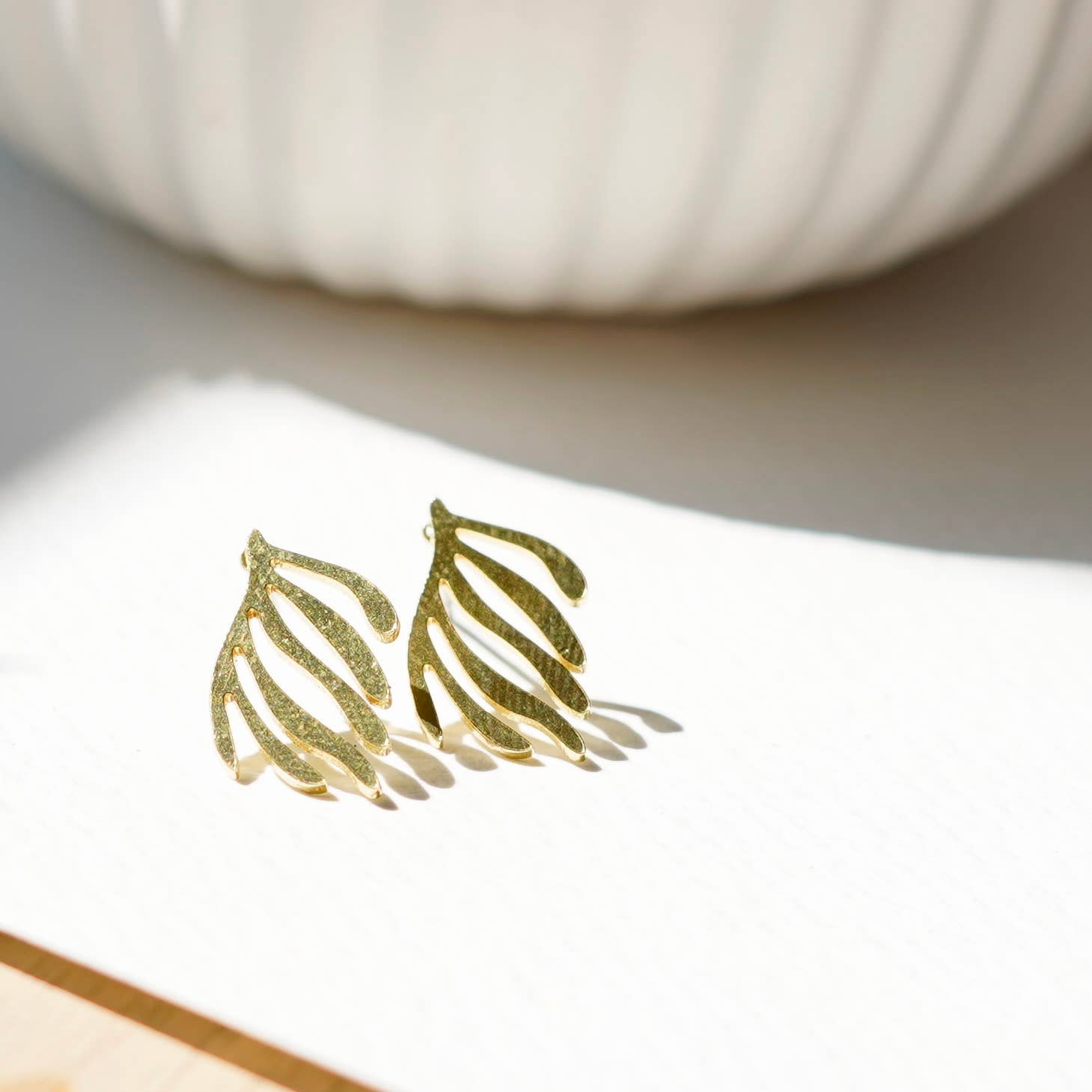 Gold leaf-shaped earrings on a white surface with a blurred pumpkin in the background