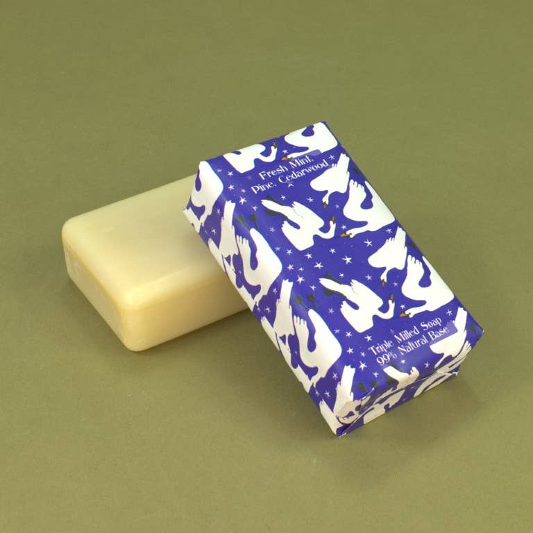 A bar of soap in a cream-colored wrapper with a blue and white unicorn pattern, placed next to an unwrapped pale yellow soap bar on a solid olive green background.