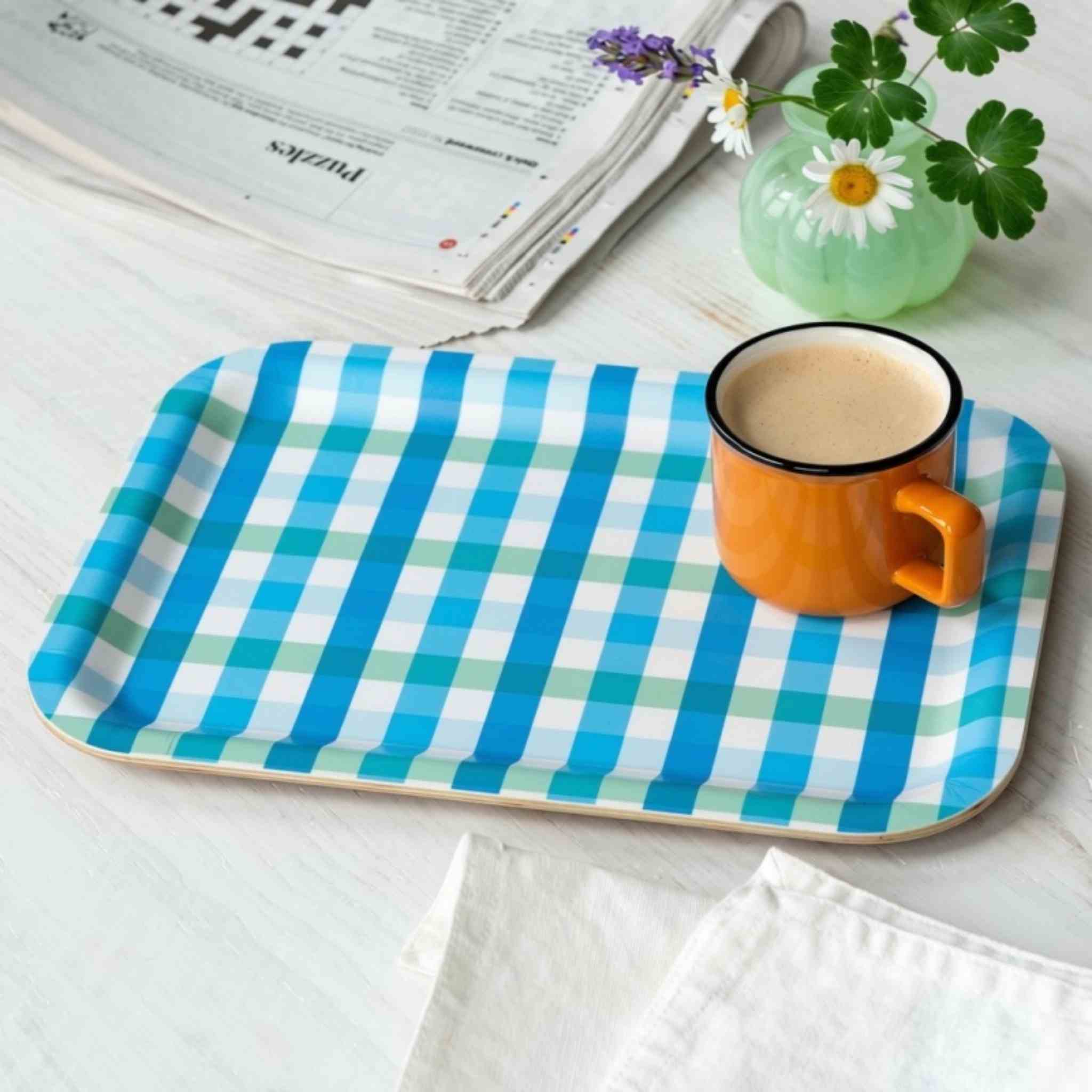 A rectangular tray with rounded corners, featuring a blue, green, and white gingham check pattern, styled on a white wooden surface with an orange mug, a small vase of flowers, and a folded newspaper.