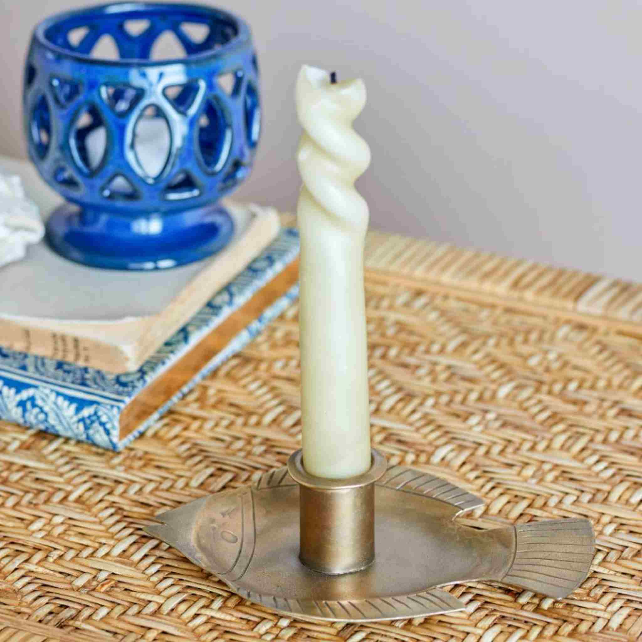 Candle in a fish-shaped brass candle holder on a woven surface with a blue candle holder in the background.