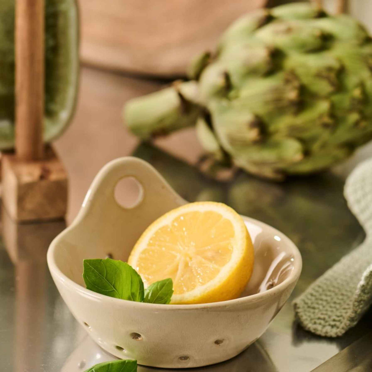 A small beige ceramic bowl with a handle and drainage holes, holding a sliced lemon and mint leaves, with a whole artichoke in the blurred background on a reflective tabletop.