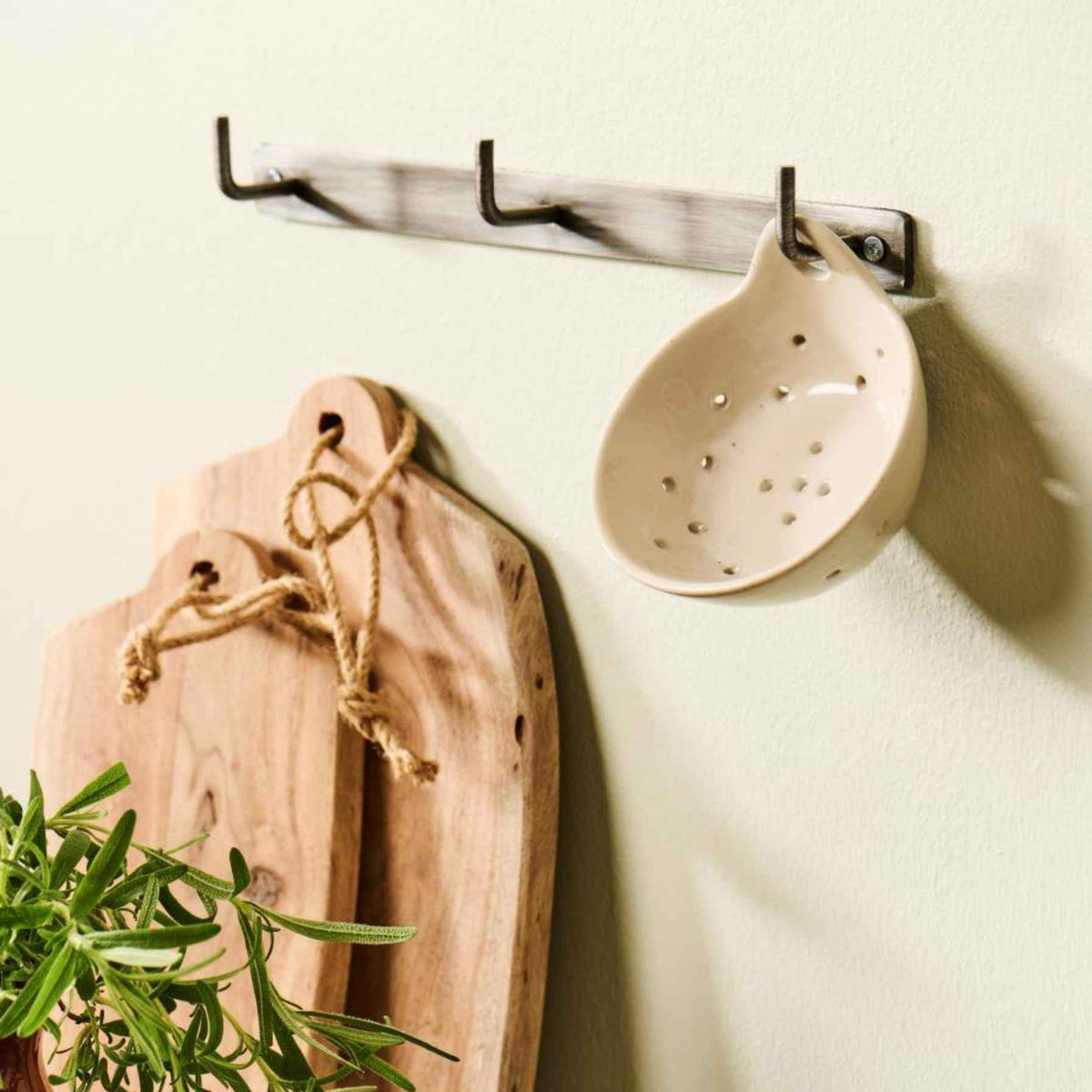 A wall-mounted hook rack with a beige ceramic colander hanging on the right and two wooden cutting boards with rope handles on the left, against a light cream wall.
