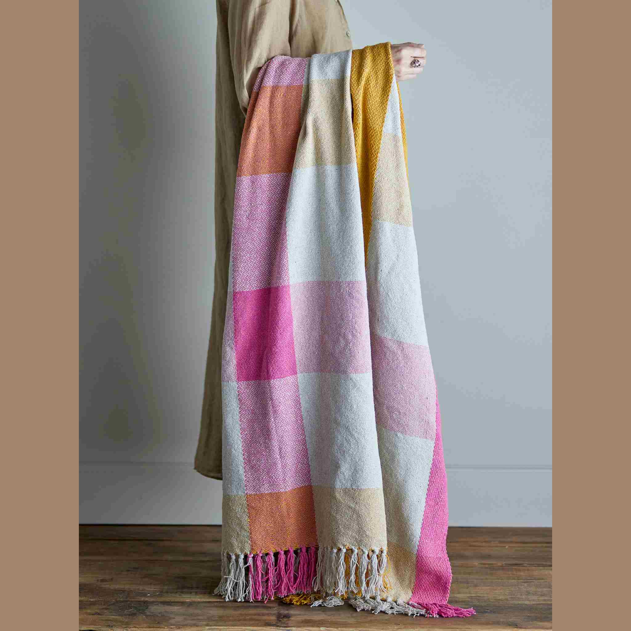 Person holding a colorful checkered blanket with pink, yellow, and white squares on a wooden floor.