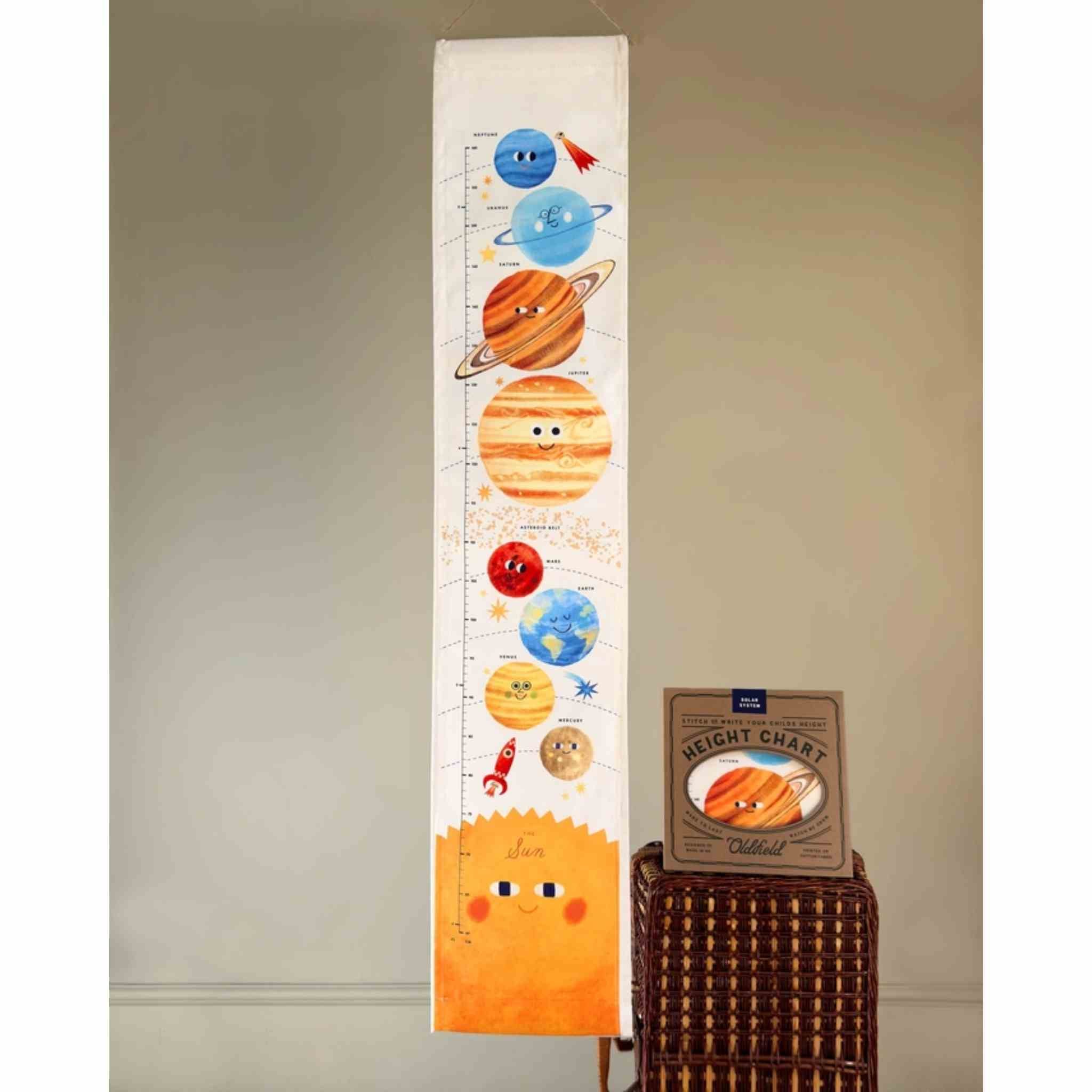 A children's height chart hanging on a wall, decorated with colorful cartoon planets and a smiling sun. A small wicker basket holding a weight chart is visible beside it against a plain beige wall.