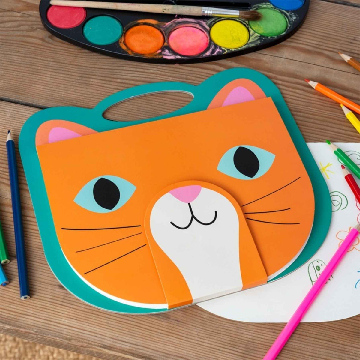 A colorful children's watercolor painting palette shaped like a cat face, displayed on a wooden table alongside drawing pencils and a watercolor paint palette.