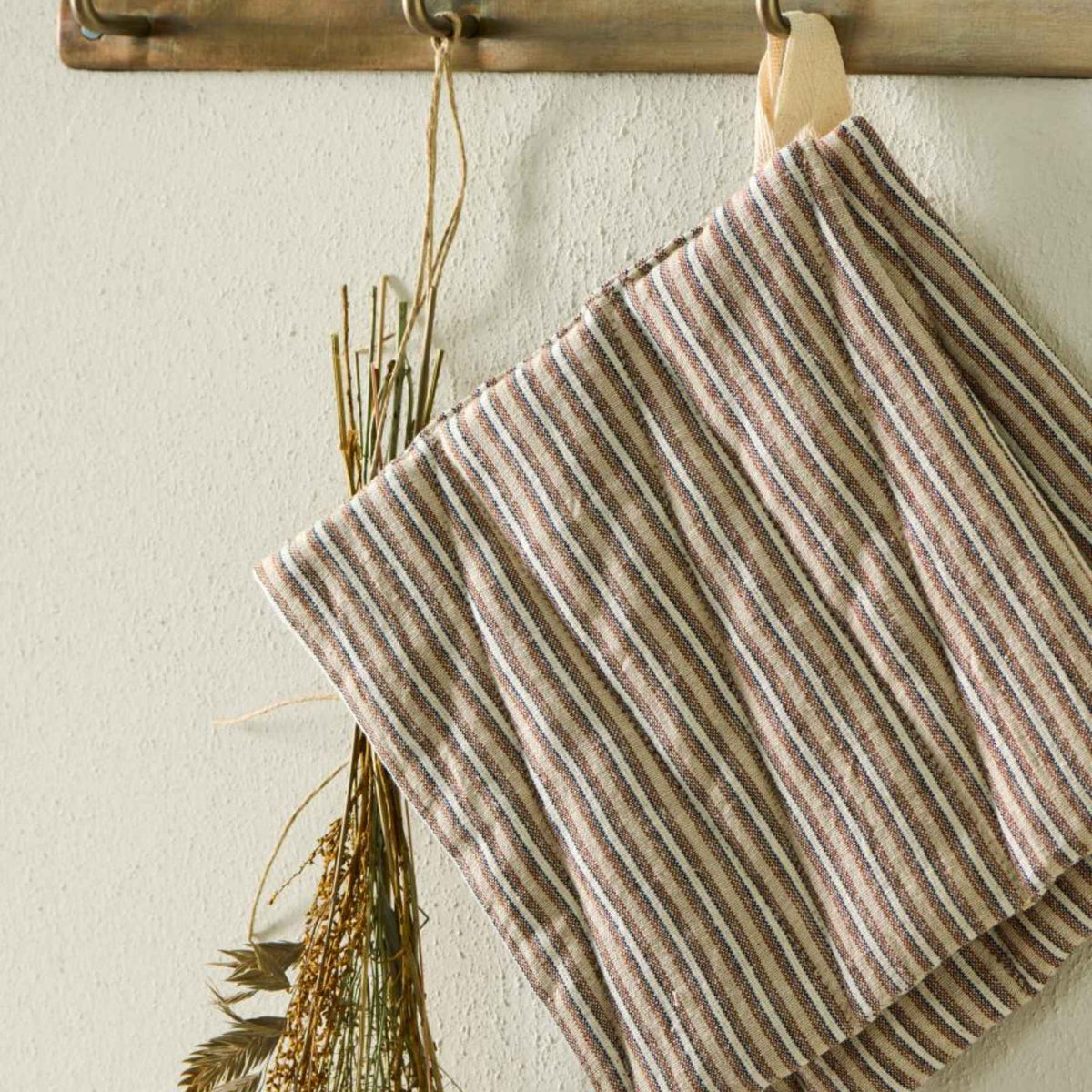 A striped square pot holder hanging from a wooden rack against a textured off-white wall, with dried grass tied to the side.