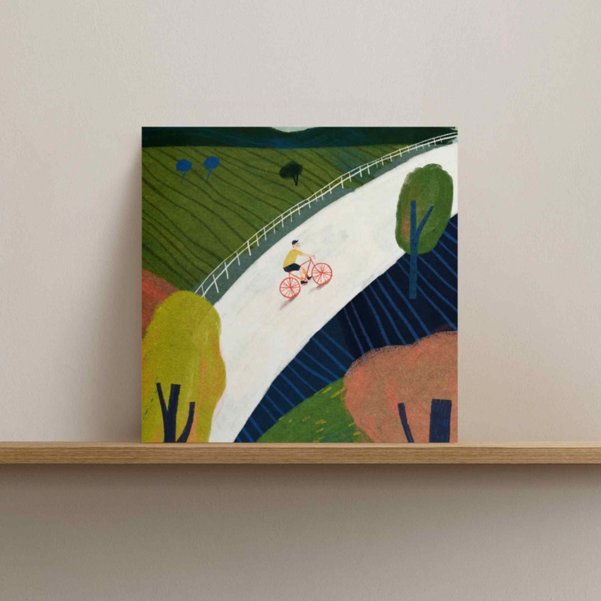 Framed artwork of a cyclist on a winding road with trees and hills.