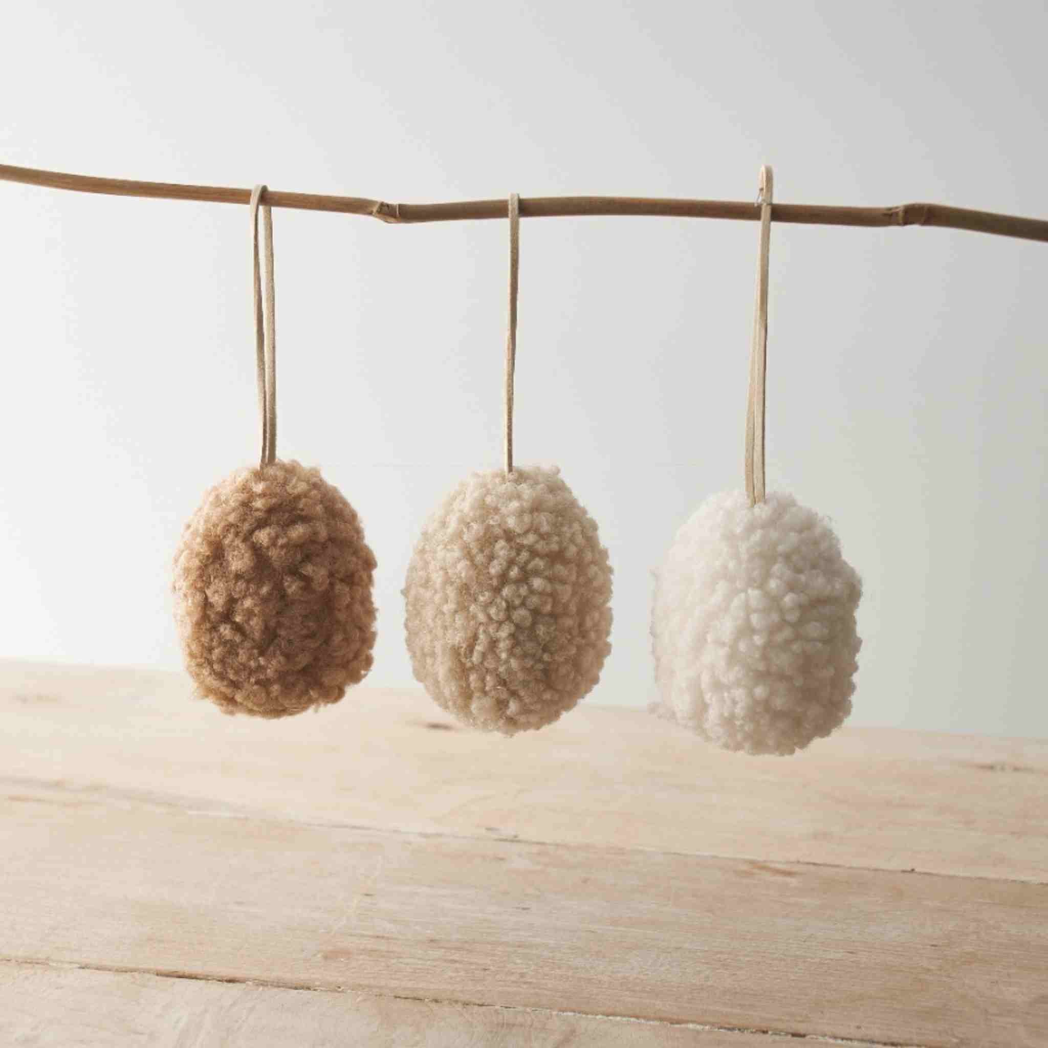 Three textured sherpa eggs hanging from a branch on a wooden surface with a light background