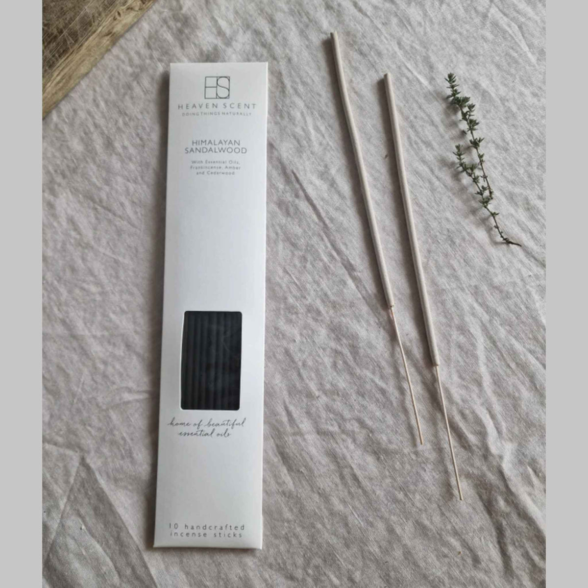 Himalayan Sandalwood incense sticks in their white packaging box, displayed on a textured light gray fabric alongside two incense sticks and a small sprig of green herbs.