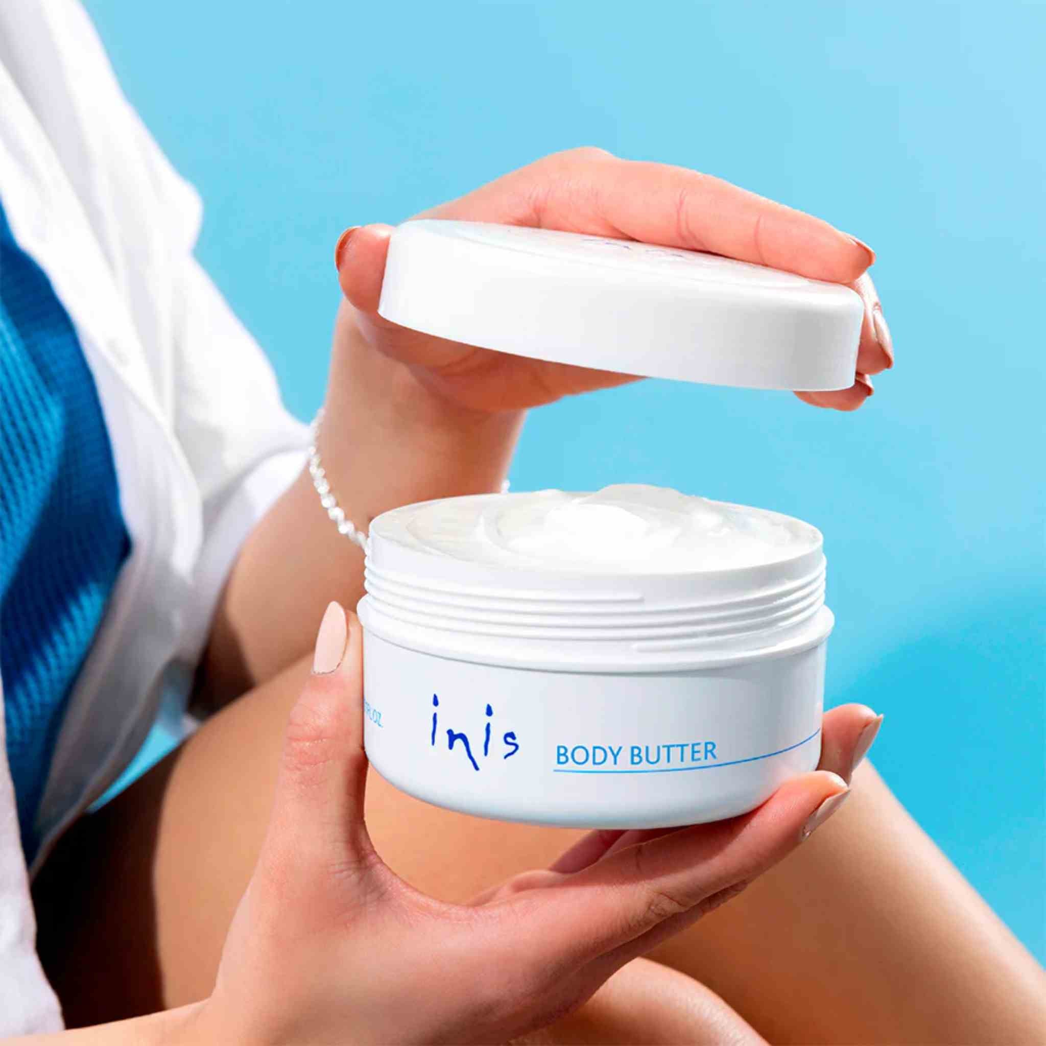 A person holds an open white jar of inis BODY BUTTER against a bright blue background. The person is wearing a white and blue shirt, and the product jar is shown with its lid off, revealing the white body butter cream inside.