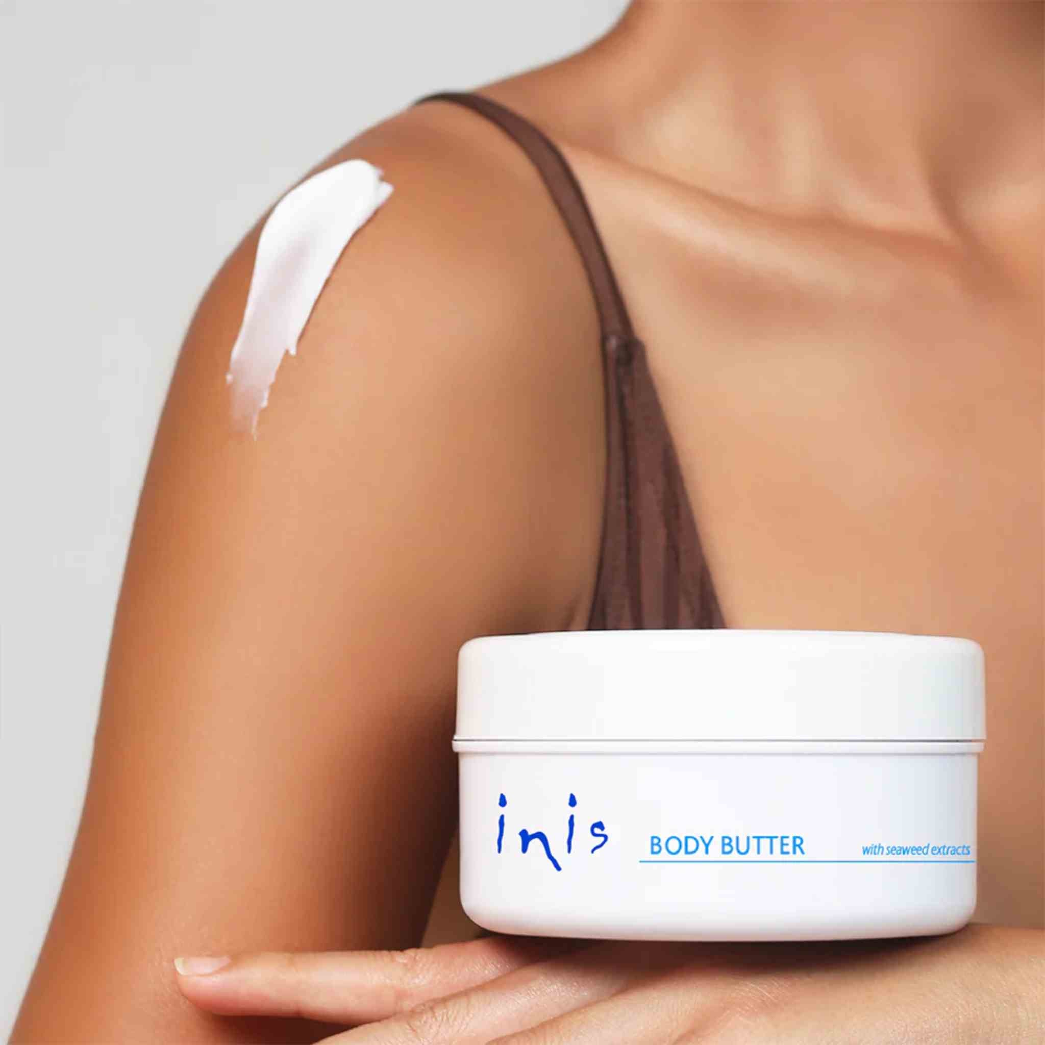 A person holding a white jar of inis Body Butter against a light gray background, with white body butter smeared on their upper arm.