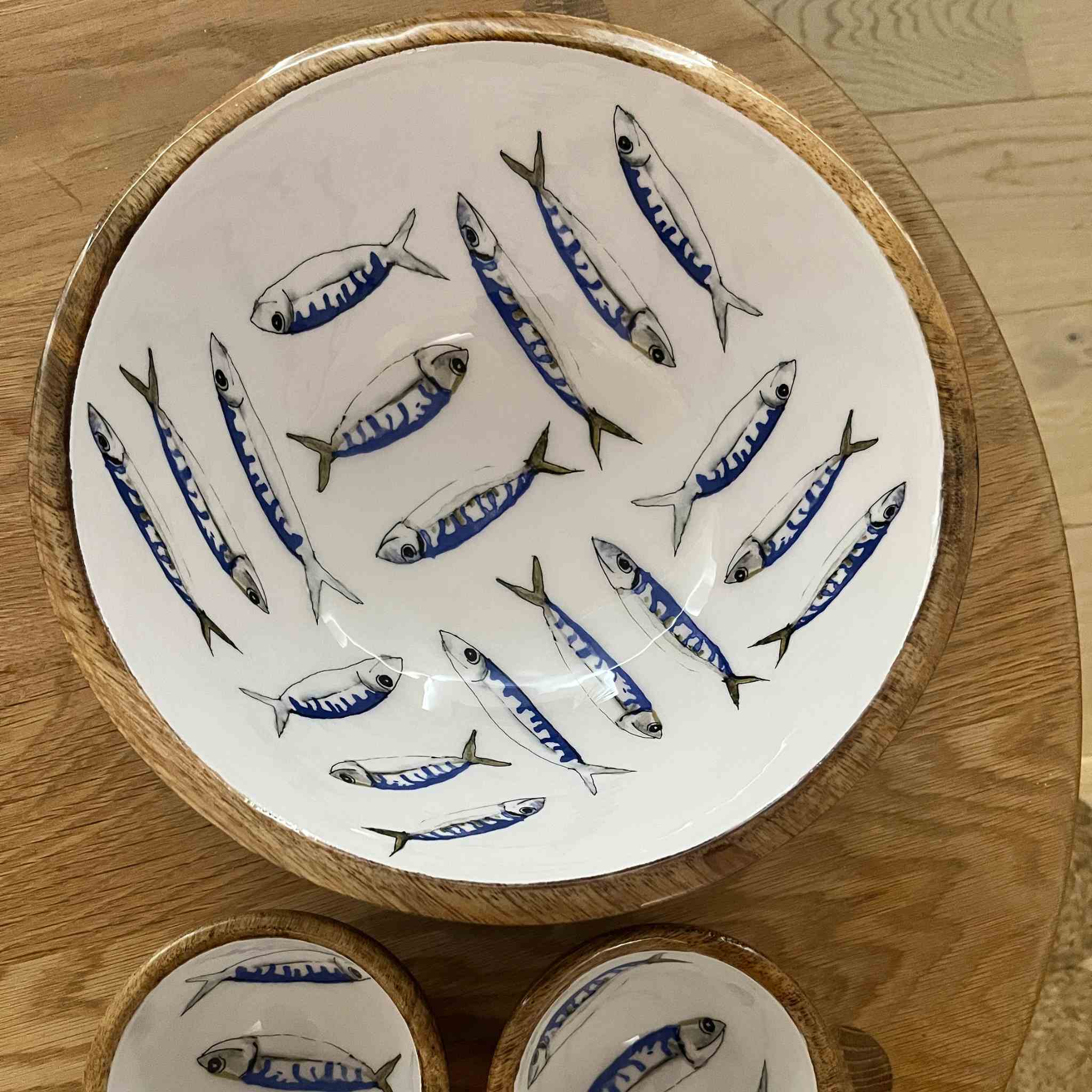 A large round ceramic bowl with a natural wood rim, decorated with painted blue and gray fish on a white background, shown on a wooden table with two matching bowls partially visible below.