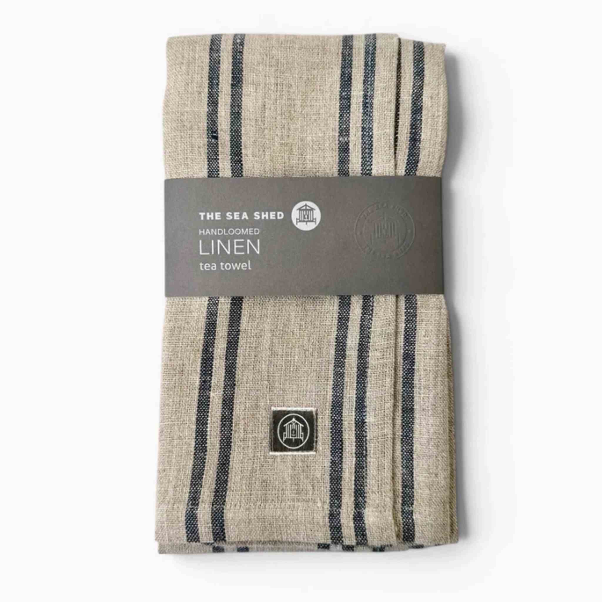A folded linen tea towel with navy vertical stripes on a beige background, wrapped in a dark gray paper band labeled 'THE SEA SHED'.