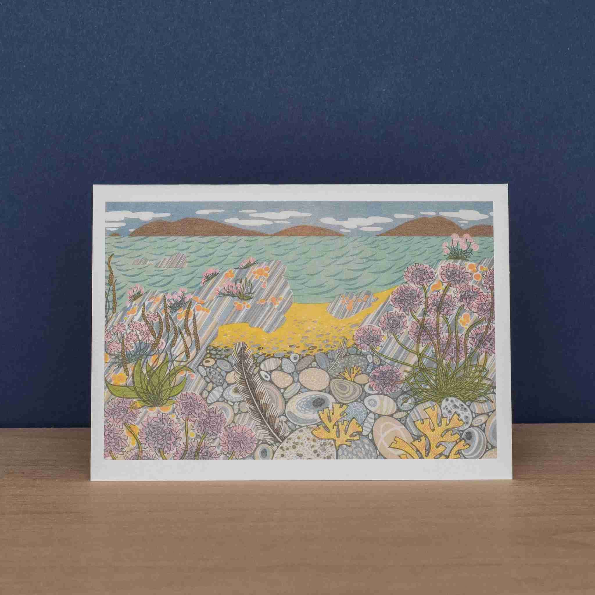 Card with a coastal scene featuring flowers, rocks, and water on a blue background