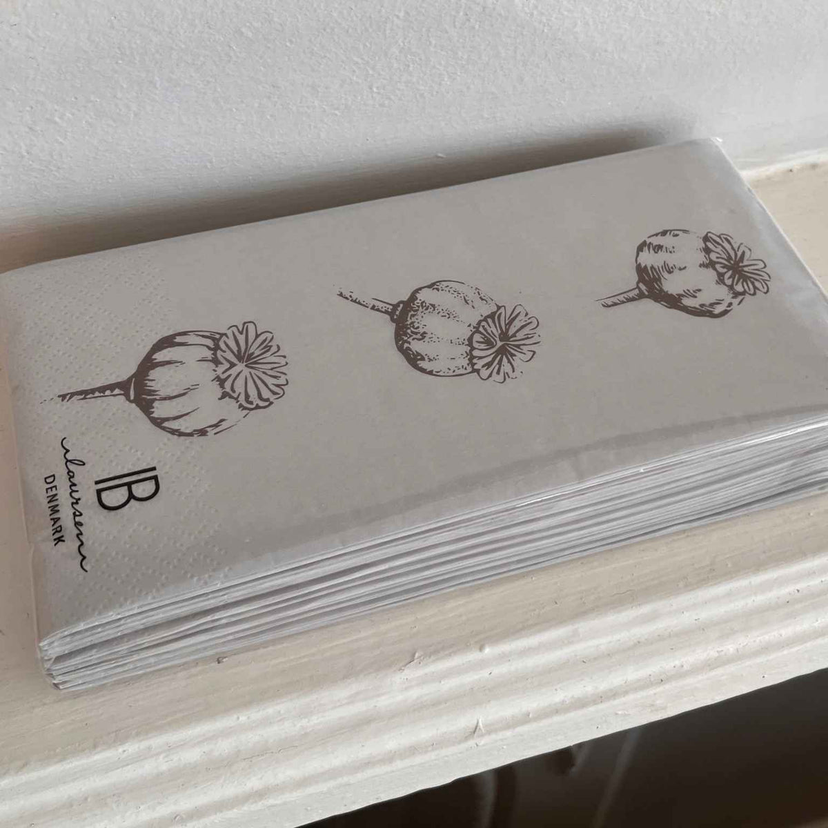 A stack of white paper napkins with embossed poppy illustrations and the brand name 'Ib Laursen' printed in black, resting on a white wooden ledge against a white wall.