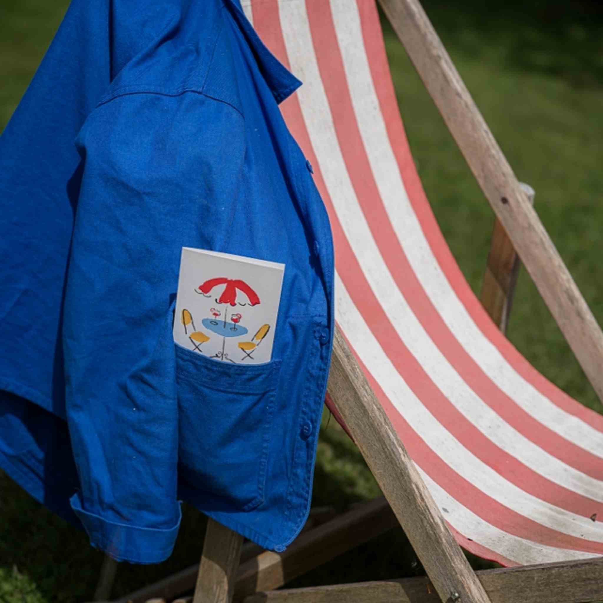 A bright blue jacket draped over a wooden deck chair outdoors, with a small card sticking up from the front pocket.