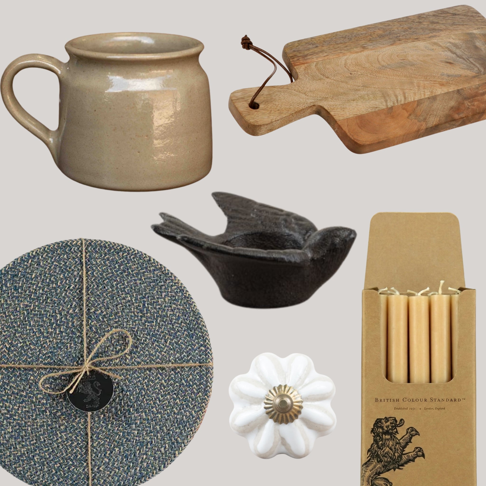 A collection of home decor and kitchen items arranged on a light gray background, including a beige ceramic pitcher, a wooden cutting board, a dark bird-shaped candle holder, a round blue textured plate wrapped in twine, a white flower-shaped cabinet knob, and a box of brass candles.