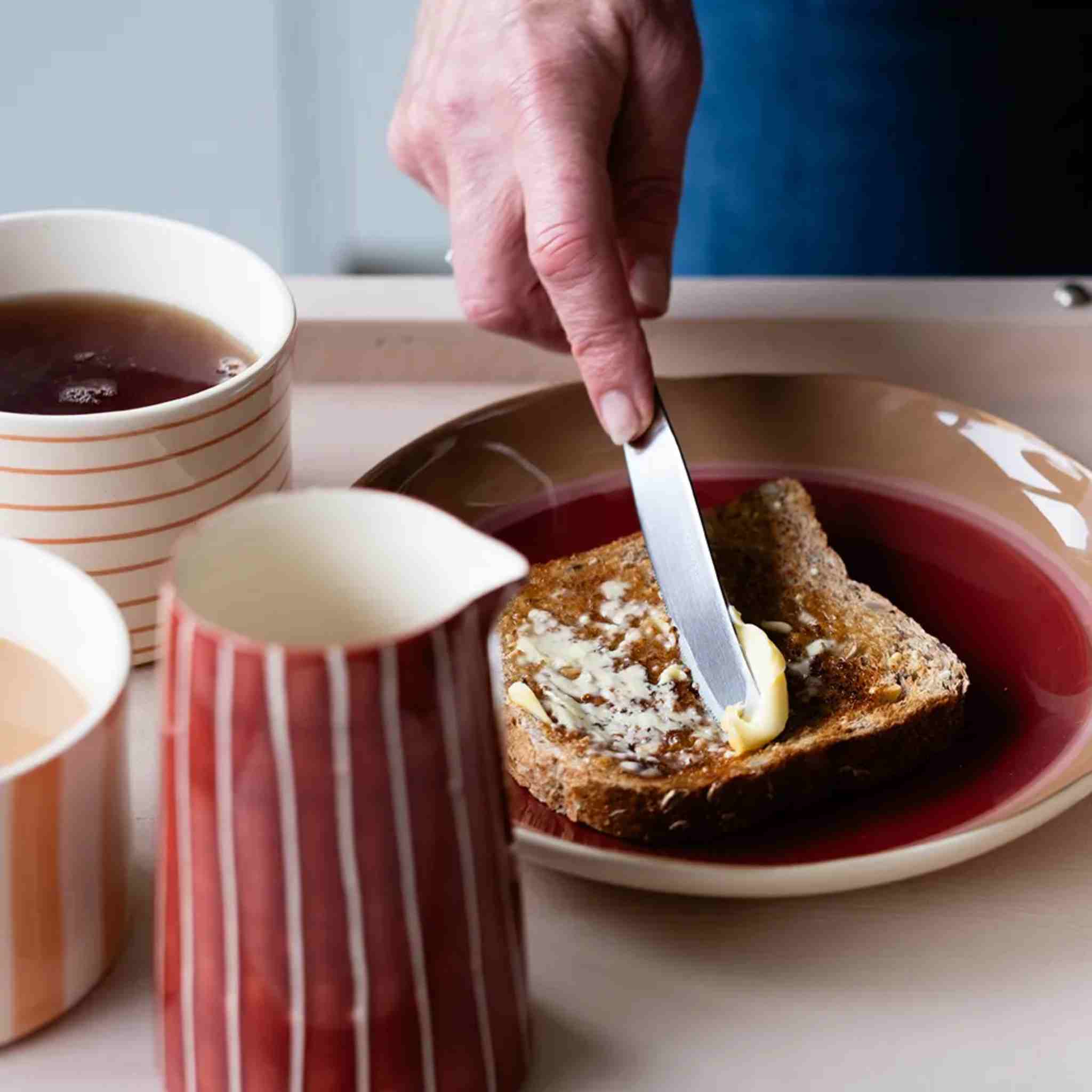 Person spreading butter on toast with a knife, next to a cup of tea and a striped milk jug.