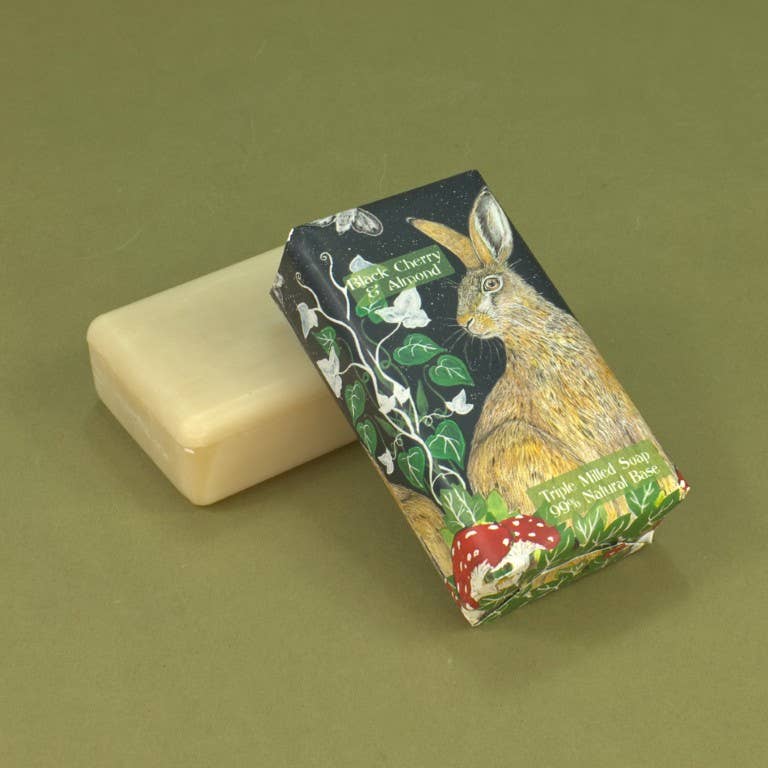 A wrapped bar of soap with a printed rabbit and floral design, placed beside an unwrapped cream-colored soap bar on an olive green background.